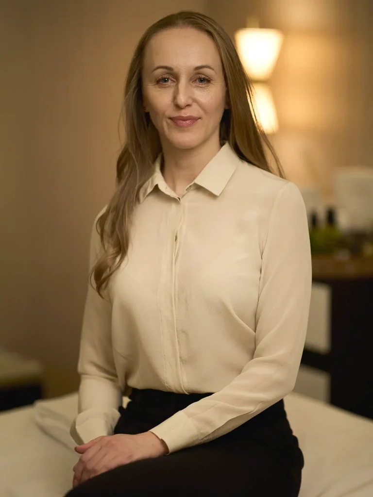 Businessporträt von Glorija in Berlin, sitzend in cremefarbener Bluse und schwarzer Hose, mit direktem Blick in die Kamera vor warm beleuchtetem Hintergrund